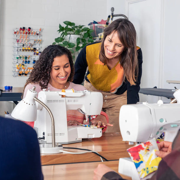A group of people gathered around sewing machines in a bright workshop or classroom setting. One woman is smiling while operating a white sewing machine, while another woman stands beside her smiling.