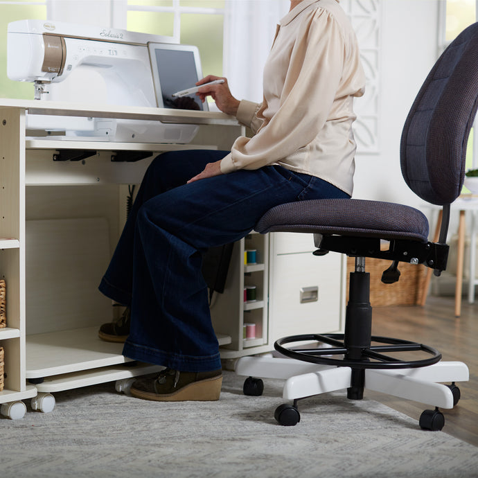 A person sits on a dark gray upholstered office chair with a white base, positioned in front of a white sewing machine and storage unit in a bright room.